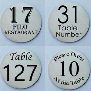 May include: Four round, silver table number signs with black text. The signs read "17 FILO RESTAURANT", "31 Table Number", "Table 127", and "Please Order 10 At the Table".