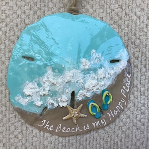May include: A blue and brown sand dollar with white painted waves and a starfish. The sand dollar has the text "The Beach is my Happy Place" written on it.