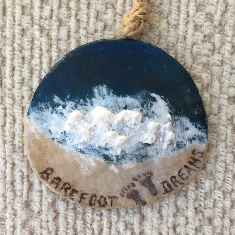 May include: Round, hand-painted ornament depicting a beach scene. The top half is dark blue, representing the ocean, with white foamy waves. The bottom half is tan, resembling sand, with the words "Barefoot Dreams" and footprints.
