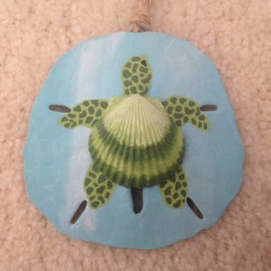 May include: A painted sand dollar with a green sea turtle design and a green seashell in the center.