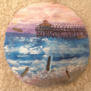 May include: A hand-painted seashell with a seascape scene. The seashell is painted with a blue ocean, white clouds, and a wooden pier with a small building on top. The sky is painted in shades of pink and purple.