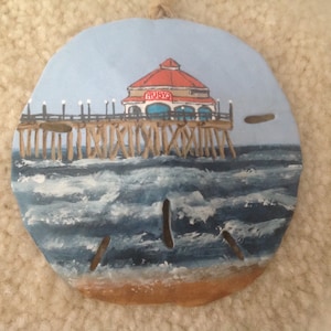 May include: Hand-painted seashell with a depiction of a pier with a red roofed building that says "Ruby's" on it. The pier is over blue water with white waves.
