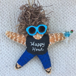 May include: A starfish ornament with brown and white shells, wearing blue jeans, a black shirt with the text "Happy Hour" and blue sunglasses. The starfish is holding a blue glass with a green olive.