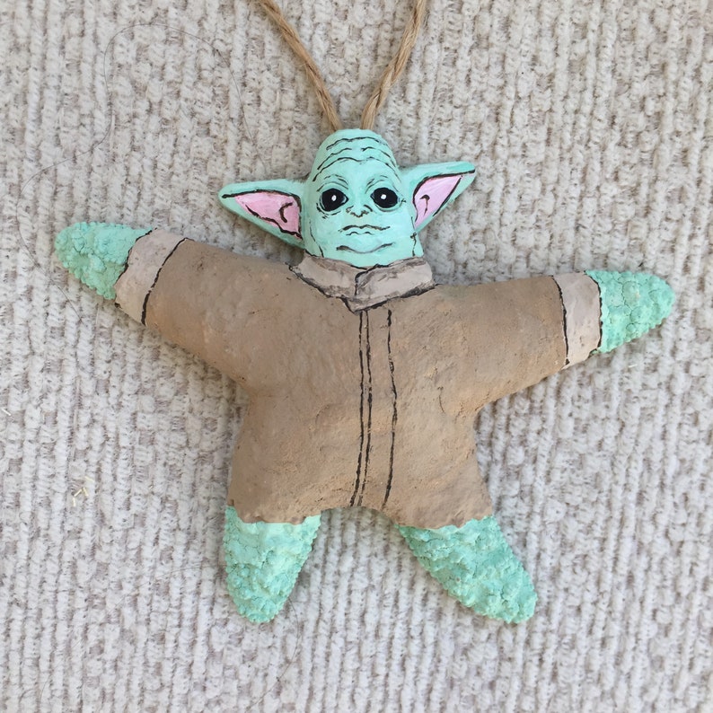 May include: A handmade ceramic ornament shaped like a star with a green and brown painted design of a popular fictional character. The ornament has a string for hanging.