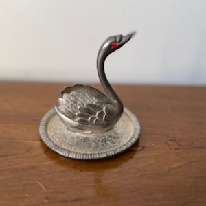 May include: A silver swan ornament with a red gem eye, standing on a decorative, round, silver plate. The swan has detailed feathering and a long, curved neck. The plate has an ornate, embossed design.