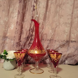 May include: A set of red and gold glassware, including a decanter and six glasses. The decanter has a tall, slender neck and a bulbous body. The glasses are small and have a stemmed base.