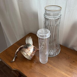 May include: A silver pitcher with an open lid, a tall clear glass vase, and a clear plastic container with a lid are on a wooden surface. The pitcher has a curved handle and ornate detailing. The vase has vertical ridges.