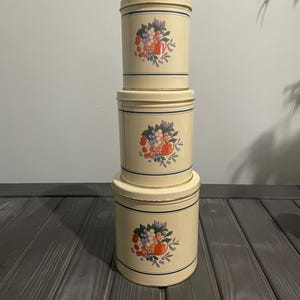 May include: Set of three vintage cream-colored metal canisters with a floral design featuring red apples, purple grapes, and blue berries.