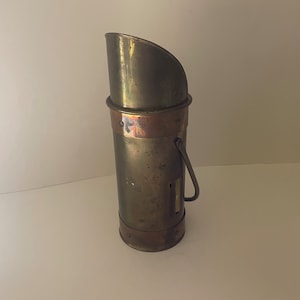 May include: A brass and copper vintage fireplace ash bucket with a handle. The bucket is slightly tarnished and has a worn finish.