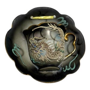 May include: A black teacup and saucer set with a dragon design. The cup and saucer have a scalloped edge and gold accents. The dragon is detailed with silver, blue, and gold. The set is likely ceramic.