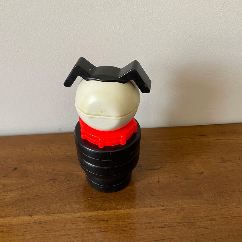 K&ouml;nnte beinhalten: Ein vintage Spielzeug aus schwarzem und wei&szlig;em Kunststoff in Form eines Hundes. Der Hund hat einen roten Kragen und schwarze Ohren. Das Spielzeug hat einen Retro-Look.