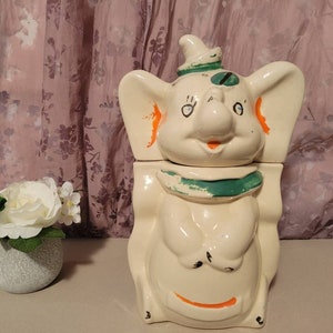 May include: A vintage ceramic elephant cookie jar with a green and orange design. The elephant has a happy expression and is wearing a green hat.