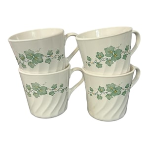 May include: Four white ceramic mugs with a green ivy leaf design. The mugs have a curved shape and a handle. The ivy design is printed around the top of the mugs. The mugs are stacked in two pairs.