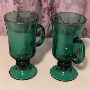 May include: Two green glass mugs with gold rims and a footed base. The mugs have a rounded shape and a handle.