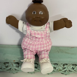 May include: A brown-skinned Cabbage Patch Kid doll wearing a white t-shirt and pink and white checkered overalls. The doll has brown hair and brown eyes.