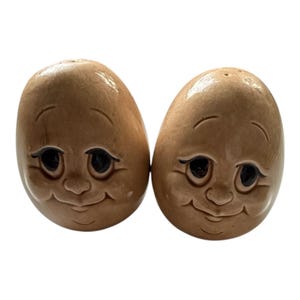 May include: A pair of vintage ceramic salt and pepper shakers. The shakers are egg-shaped with a light brown glaze and feature smiling faces with black eyes and delicate facial details. The shakers are designed to resemble cartoon characters.