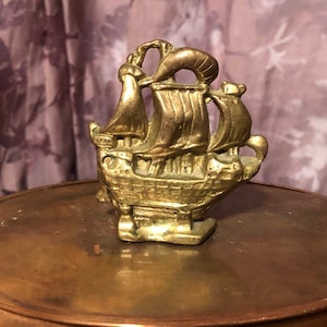 May include: A gold-coloured metal ship figurine with intricate details. The ship has a tall mast and sails, and a detailed hull. The figurine is sitting on a copper surface.