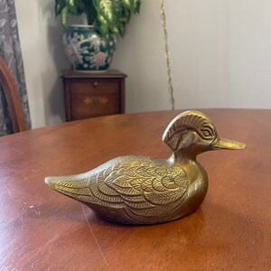 Puede incluir: Una figurita de pato de lat&oacute;n de color dorado con detalles intrincados en sus plumas. El pato est&aacute; sentado sobre una superficie de madera.