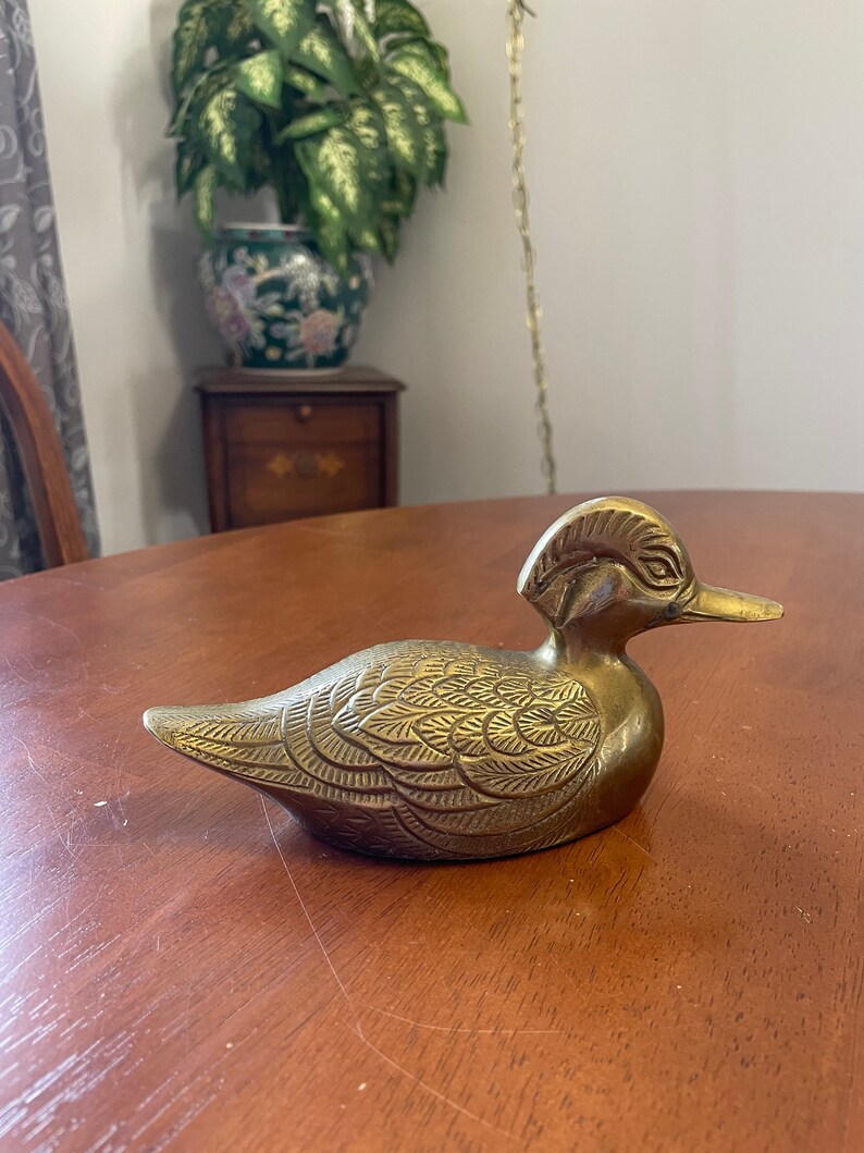 Puede incluir: Una figurita de pato de lat&oacute;n de color dorado con detalles intrincados en sus plumas. El pato est&aacute; sentado sobre una superficie de madera.