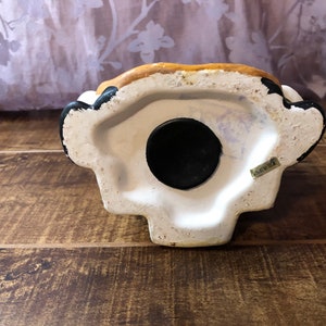May include: The bottom of a ceramic piggy bank with a black rubber stopper. The bank is painted white with brown and black accents. The words "Made in China" are printed on the bottom.