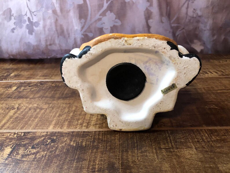 May include: The bottom of a ceramic piggy bank with a black rubber stopper. The bank is painted white with brown and black accents. The words "Made in China" are printed on the bottom.