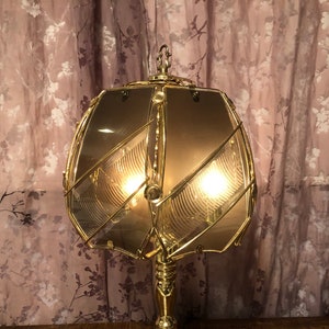 May include: A gold-toned table lamp with a geometric, clear glass shade. The lamp has a decorative, ornate design and is lit with two light bulbs.