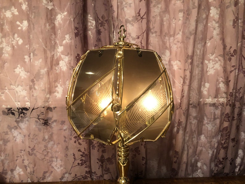 May include: A gold-toned table lamp with a geometric, clear glass shade. The lamp has a decorative, ornate design and is lit with two light bulbs.