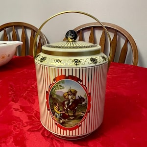 Peut inclure: Une jarre à biscuits en céramique blanche et dorée avec un motif rayé rouge et blanc. La jarre a une poignée dorée et un couvercle avec un bouton noir. La jarre présente une scène peinte d'une personne montant un âne.