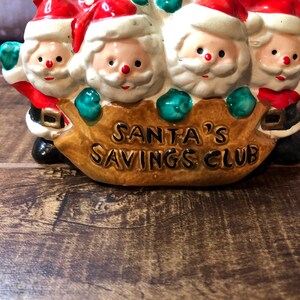 May include: A ceramic piggy bank shaped like four Santa Clauses carrying a brown sack with the words "Santa's Savings Club" on it.