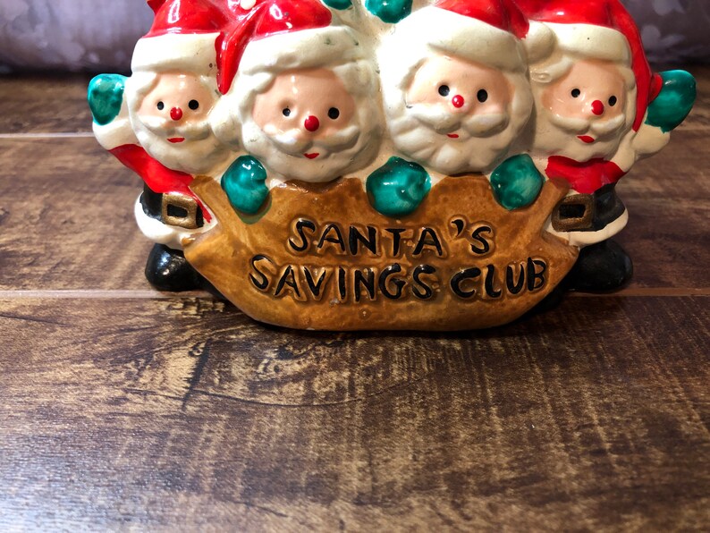 May include: A ceramic piggy bank shaped like four Santa Clauses carrying a brown sack with the words "Santa's Savings Club" on it.
