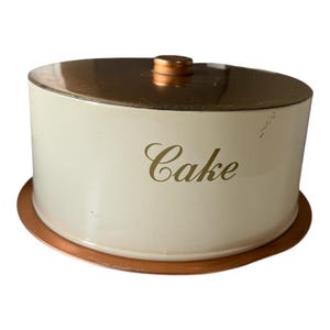 Peut inclure: Une boîte à gâteau vintage avec un couvercle et une base dorés. Le corps blanc cassé du récipient porte le mot "Cake" en lettres dorées. Le récipient est rond avec un petit bouton sur le dessus.