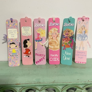 May include: Six colourful bookmarks featuring different images and text. The bookmarks are pink, blue and white with images of Barbie dolls, Peanuts characters, and text that reads "Girls just want to have fun." "Reading makes you beautiful!" "Party Party Party" "Really Cool" "Nice Goin'" and "Dreams can come true!"