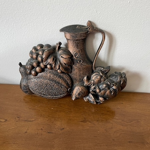 May include: A wall decoration made of metal, featuring a pitcher, grapes, and other fruits. The piece is painted in a bronze colour.