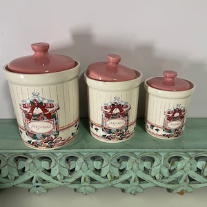 May include: Set of three white ceramic canisters with pink lids and floral designs. Each canister has the word "Potpourri" written on it.
