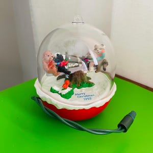 Peut inclure: Une boule de Noël transparente avec une base rouge et un bord festonné blanc. À l'intérieur, des figurines d'ours sont rassemblées autour d'une souche d'arbre. L'ornement a un cordon d'alimentation vert et le texte "Merry Christmas 1992".