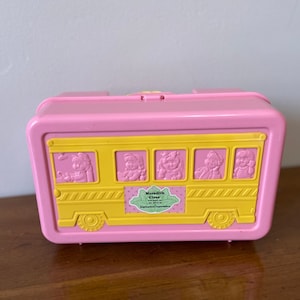 May include: A pink plastic lunchbox with a yellow school bus design. The bus has windows with cartoon faces inside and the text "Meredith Ciree" on the side.