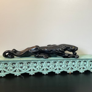 May include: A black panther figurine with a gold eye, lying on a light blue shelf with a carved design.