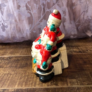 May include: A ceramic Christmas figurine of a stack of Santas, each wearing a red suit with white trim, a black hat, and green mittens. The Santas are standing on a white platform with brown accents.