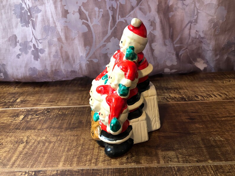 May include: A ceramic Christmas figurine of a stack of Santas, each wearing a red suit with white trim, a black hat, and green mittens. The Santas are standing on a white platform with brown accents.