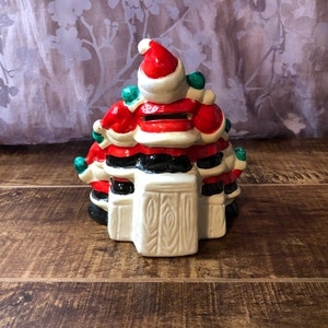 May include: A ceramic Christmas tree shaped piggy bank with a Santa Claus design. The tree is decorated with red, white, and green. The Santa Claus is wearing a red hat and a white beard.