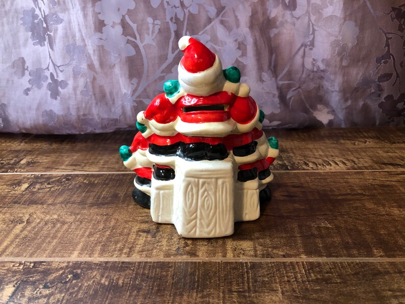 May include: A ceramic Christmas tree shaped piggy bank with a Santa Claus design. The tree is decorated with red, white, and green. The Santa Claus is wearing a red hat and a white beard.