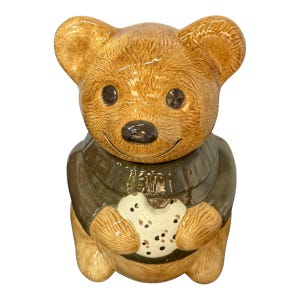 May include: A ceramic teddy bear cookie jar. The bear is tan with a dark green jacket and is holding a white biscuit with dark brown spots. The bear has black eyes and a black nose. A decorative kitchen item.