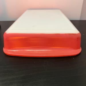 Puede incluir: Un recipiente rectangular de dos tonos con una tapa blanca y una base roja vibrante. El recipiente tiene bordes redondeados y parece estar hecho de plástico. El diseño es simple y funcional, adecuado para el almacenamiento o la organización.