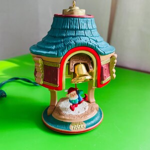 May include: A Christmas ornament featuring a bell-shaped structure with a teal roof and red accents. A small figure of Santa Claus is suspended from the bell. The base of the ornament is marked with the year "1993".