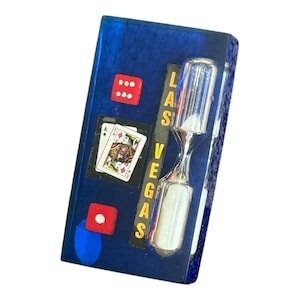 May include: A rectangular, blue decorative item featuring a silver hourglass, dice, and playing cards. The word "LAS VEGAS" is printed vertically in gold. The playing card shows the Ace of Spades.