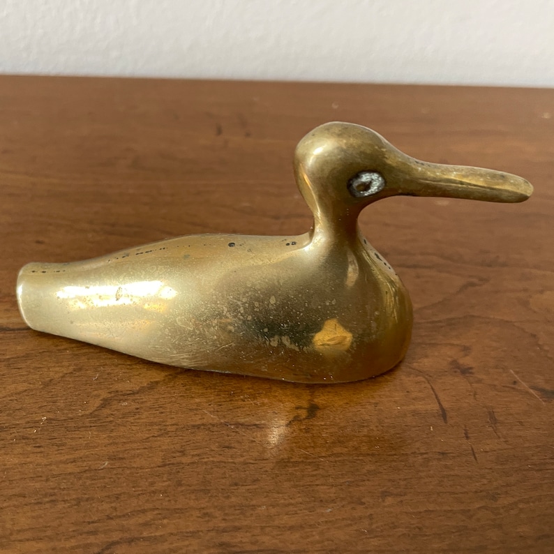 May include: A small, brass duck figurine with a smooth, shiny finish. The duck has a long, slender beak and a round body. The eyes are inlaid with white dots.