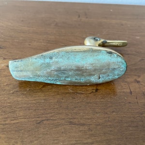 May include: A brass duck figurine with a green patina. The duck is standing on a wooden surface.