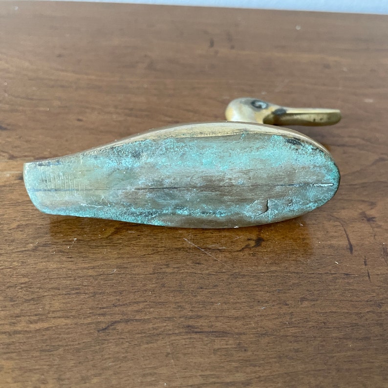 May include: A brass duck figurine with a green patina. The duck is standing on a wooden surface.