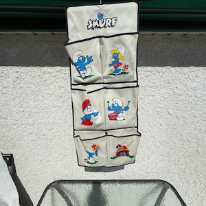 May include: A vintage Smurf-themed hanging organizer with multiple pockets. The off-white fabric features blue trim and illustrations of various Smurf characters. The word "SMURF" is printed at the top.