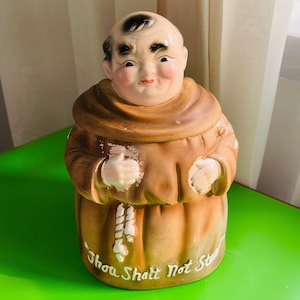 May include: A ceramic figurine of a monk in a brown robe with a smiling face and black hair. The figurine has the text "Thou Shalt Not Steal" at the base. The monk's face is painted with rosy cheeks and a small smile.
